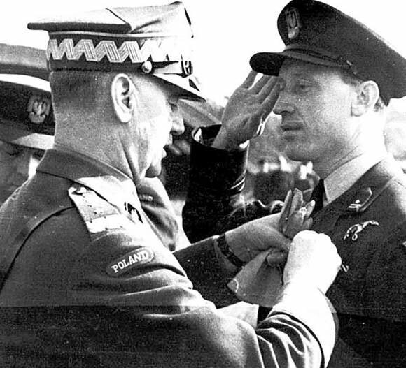 Zdjęcie przedstawia młodego mężczyznę w mundurze polskiego lotnika, który salutuje Generałowi Sikorskiemu, podczas gdy ten przypina mu do piersi odznaczenie.