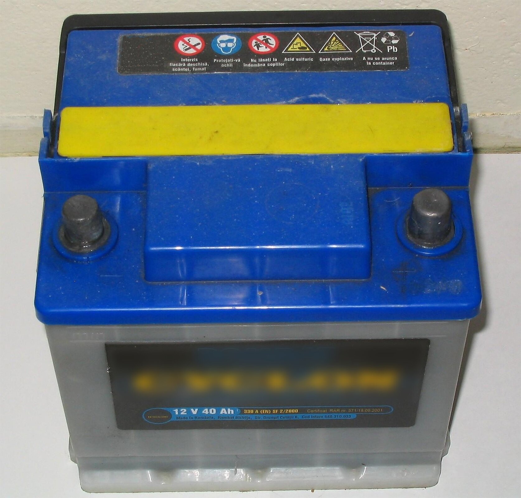 Zdjęcie przedstawia samochodowy akumulator ołowiowy pokazany z przodu i z góry. Obudowa jest szara, a pokrywa niebieska z żółtą rączką. Posiada on dwie grube metalowe elektrody po prawej i lewej stronie. Na etykiecie z przodu znajduje się nazwa marki i parametry: napięcie dwanaście woltów, pojemność czterdzieści amperogodzin.