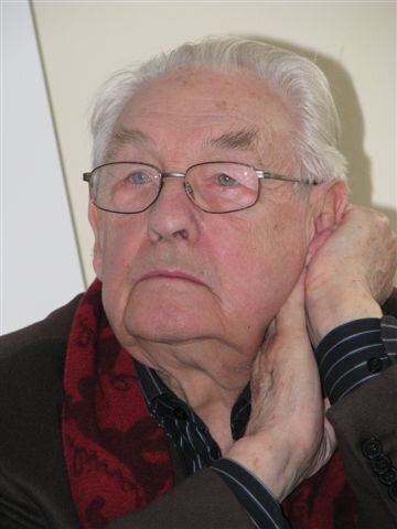 Ilustracja przedstawia starszego mężczyznę. Postać ukazuje lewy półprofil. Czoło mężczyzny jest wysokie i pomarszczone, brwi są przerzedzone. Mężczyzna ma opadające powieki. Patrzy przed siebie. Mężczyzna ma duży nos i wąskie usta. Nosi okulary w wąskiej oprawie. Człowiek podpiera rękami głowę. Mężczyzna jest ubrany w prążkowaną koszulę i marynarkę. Na szyi ma wzorzysty, czerwony szal.