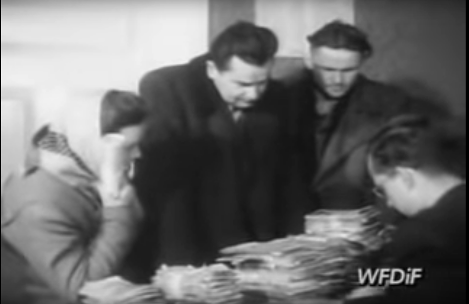 Fotografia przedstawia dwóch mężczyzn i kobietę stojących przy ladzie w banku, gdzie leży kilka kupek pieniędzy. Po drugiej stronie siedzi pracownik banku i liczy pieniądze.