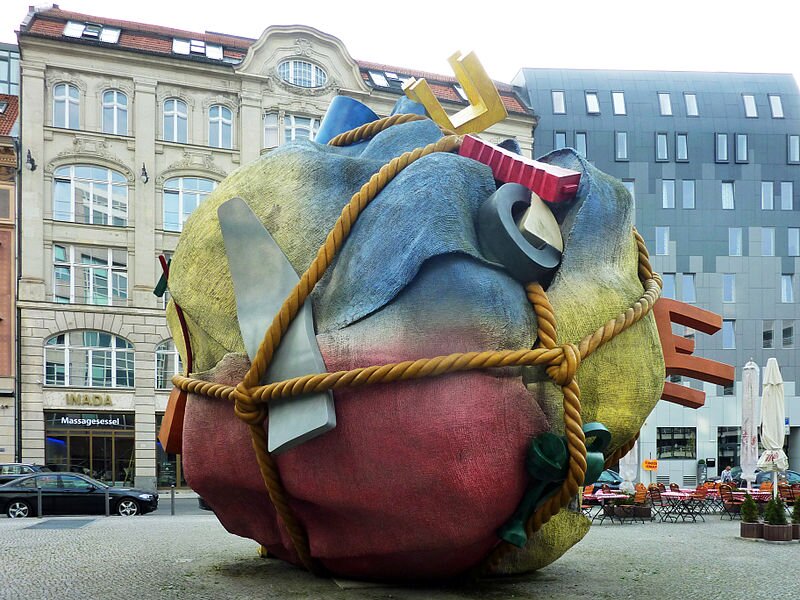 Na zdjęciu znajduje się jedenastometrowa kulista rzeźba. Wokół owinięta jest liną z supełkami. Z lewej strony za liną znajdują się kawałki stali nierdzewnej. Po prawej stronie są umocowane kawałki pianki poliuretanowej w różnych kolorach i kształtach. Rzeźba stoi na środku brukowanego placu w pobliżu jezdni, gdzie zostało zaparkowane czarne auto. Po prawej stronie znajduje się kawiarniany ogródek, gdzie są ustawione stoliki z dużymi białymi parasolami. Przy stolikach są ustawione krzesła. Ogródek zdobią małe tuje w brązowych drewnianych skrzynkach. Za rzeźbą są widoczne dwa budynki. Budowla stojąca po lewej stronie to kamienica, a po prawej stoi nowoczesny szklany budynek.