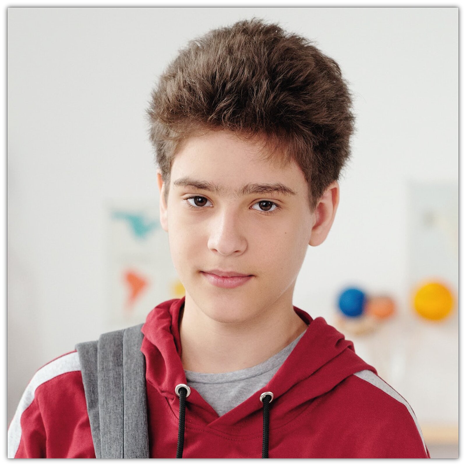 Na zdjęciu widoczny jest młody chłopak w czerwonej bluzie. Chłopak stoi w klasie lekcyjnej. Na jednym ramieniu niesie plecak. Ma spokojny wyraz twarzy. 