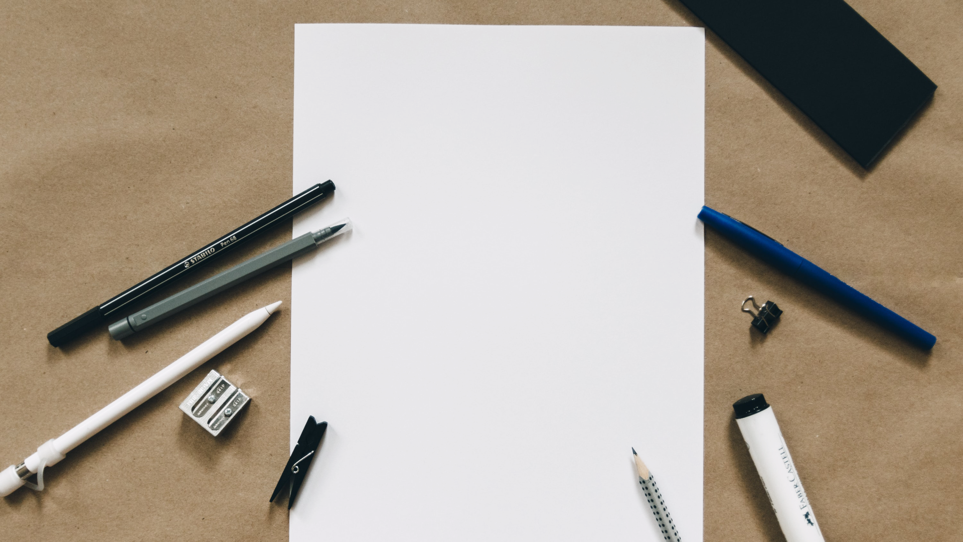 Zdjęcie przedstawia kartkę, która lezy na stole. Zdjęcie zrobione jest z góry. Obok pustej, niezapisanej kartki leżą cztery różne długopisy, ołówek, pisak, temperówka oraz dwa spinacze.