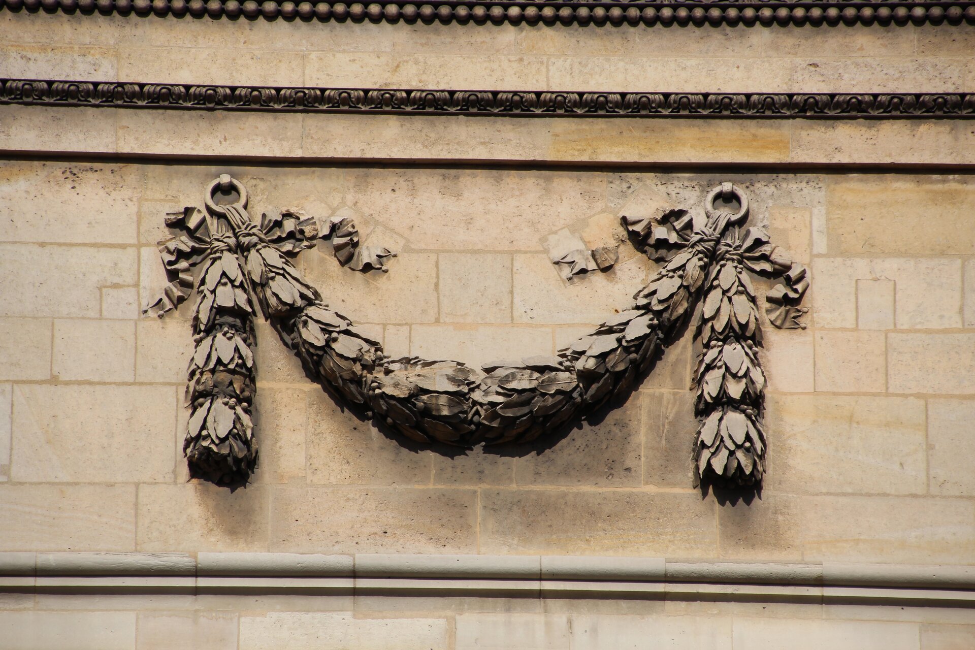 Fotografia przedstawia ornament, który umieszczony został na ścianie budynku. Ma on kształt litery M i składa się z wymodelowanych liści oraz wstążek. Ornament wisi na tle jasnej ściany.