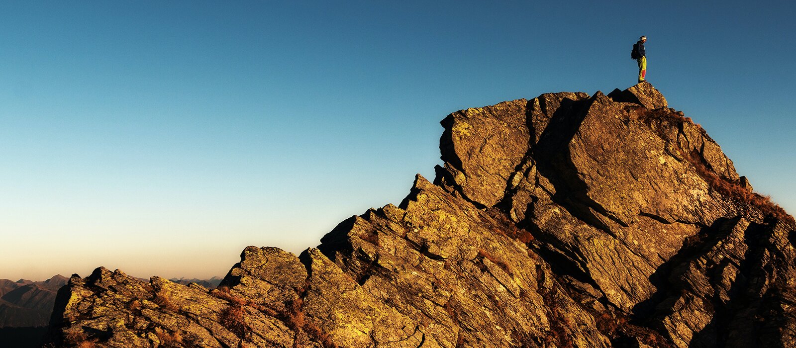 Zdjęcie przedstawia postać człowieka stojącego na szczycie skalistej góry. Widoczne są brązowe skały, oświetlane promieniami słońca. W najwyższym punkcie widoczny jest stojący człowiek z plecakiem, zwrócony w prawo. W tle widoczne jest niebieskie niebo. 
