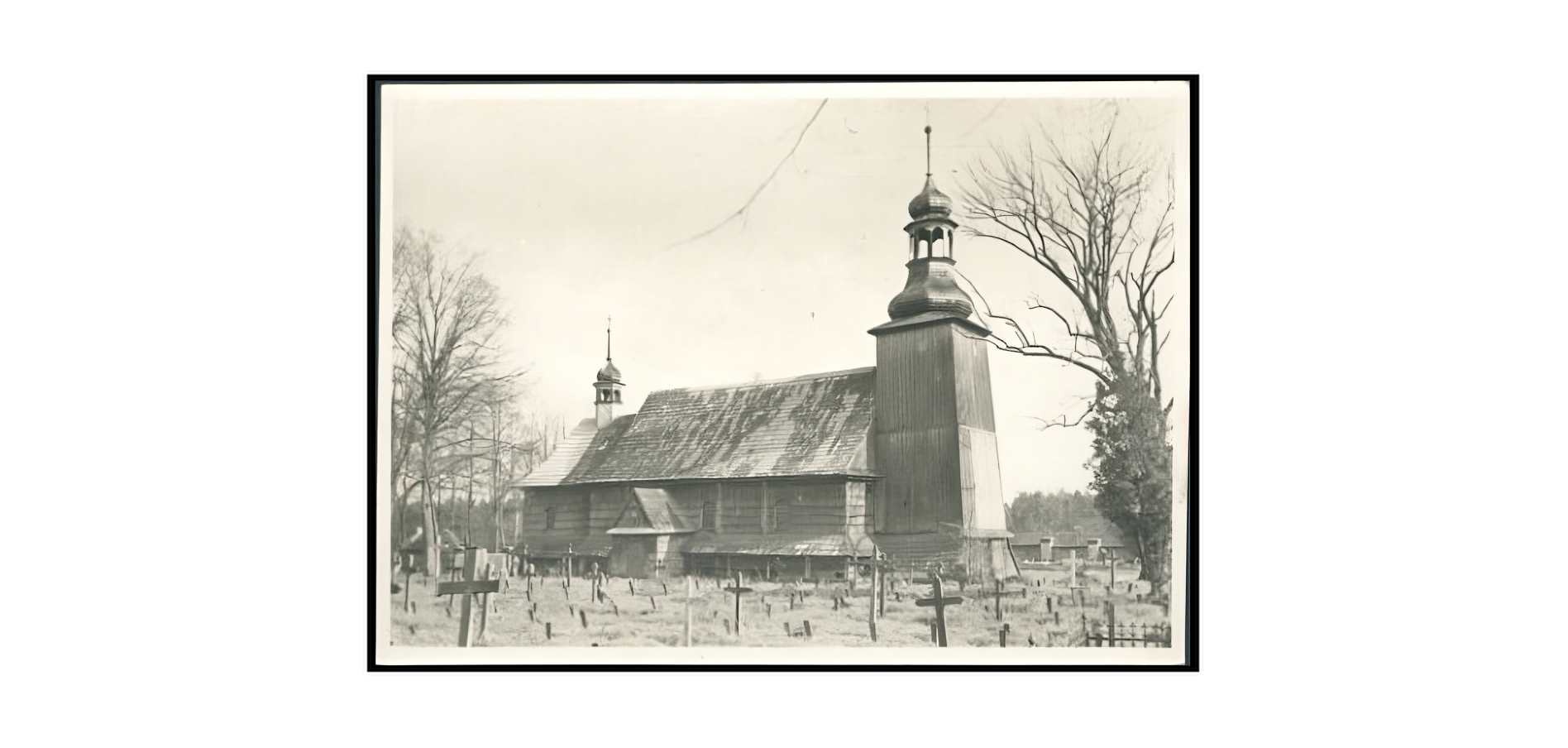 Zdjęcie archiwalne przedstawia budynek kościoła w Koszęcinie. Drewniana konstrukcja o dwuspadowym dachu, z wieżyczką dzwonną od strony kruchty (po lewej stronie fotografii), sygnaturką nad amboną (po prawej) i skrzydłem z bocznym wejściem znajduje się w centrum ilustracji. Na szczycie obu wież dzwonnych metalowe kopuły w stylu barokowym. Na pierwszym planie krzyże cmentarne, rozstawione nieregularnie na polu cmentarza. Po obu stronach kościoła na fotografii widoczne bezlistne drzewa. 