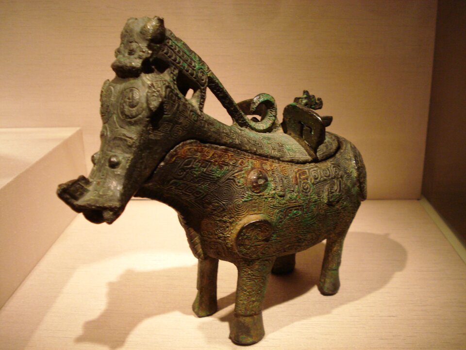 Zdjęcie przedstawia eksponat muzealny w postaci naczynia w kształcie czworonożnego zwierzęcia na którego grzbiecie siedzi jaszczurka z zawiniętym ogonem.