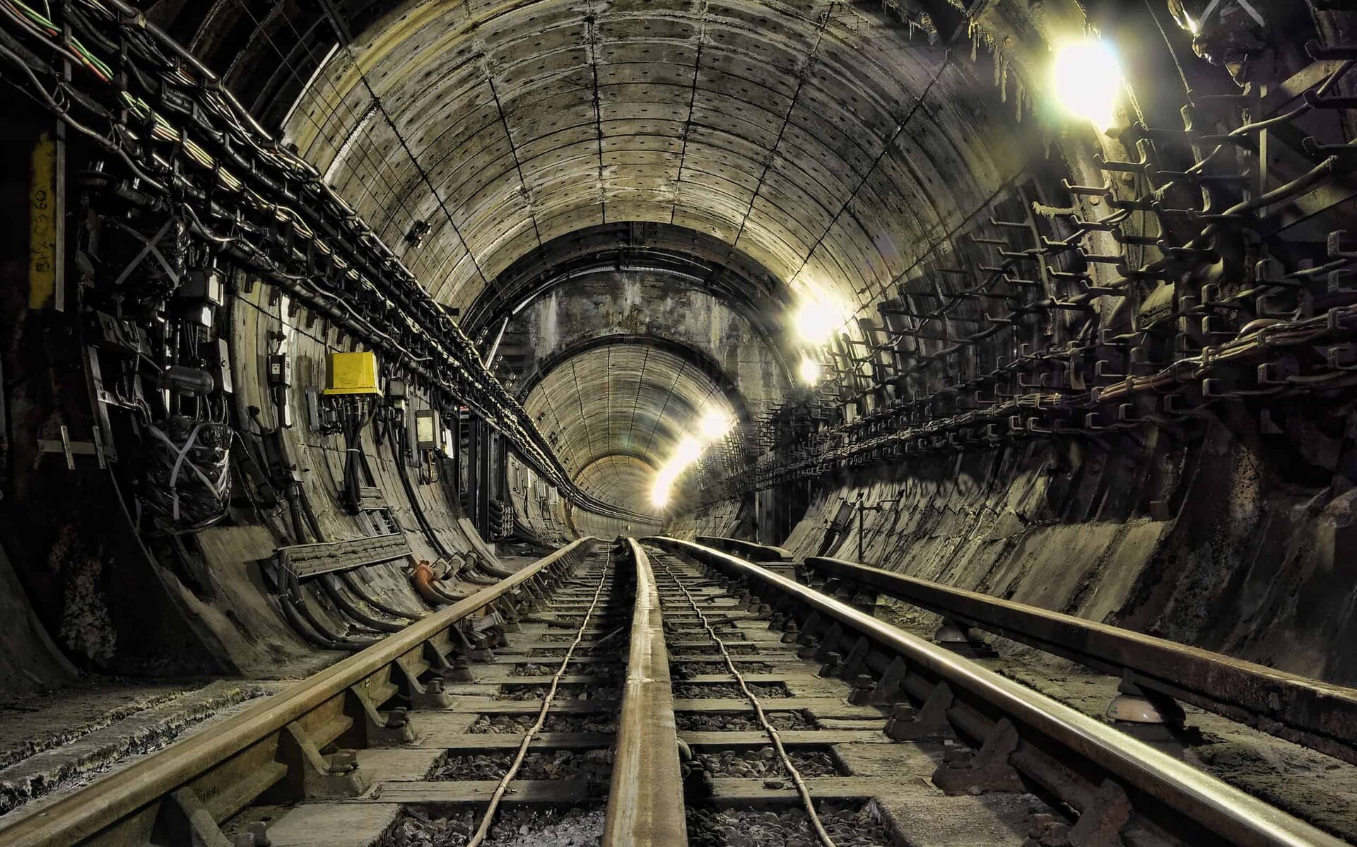 Zdjęcie przedstawia wnętrze betonowego tunelu kolejowego. Na ścianach widoczne są liczne kable i instalacje, co wskazuje na złożoną infrastrukturę zapewniającą oświetlenie i inne potrzeby tunelu. Obiekt jest dobrze oświetlony, co zapewnia widoczność i bezpieczeństwo w jego użytkowaniu.