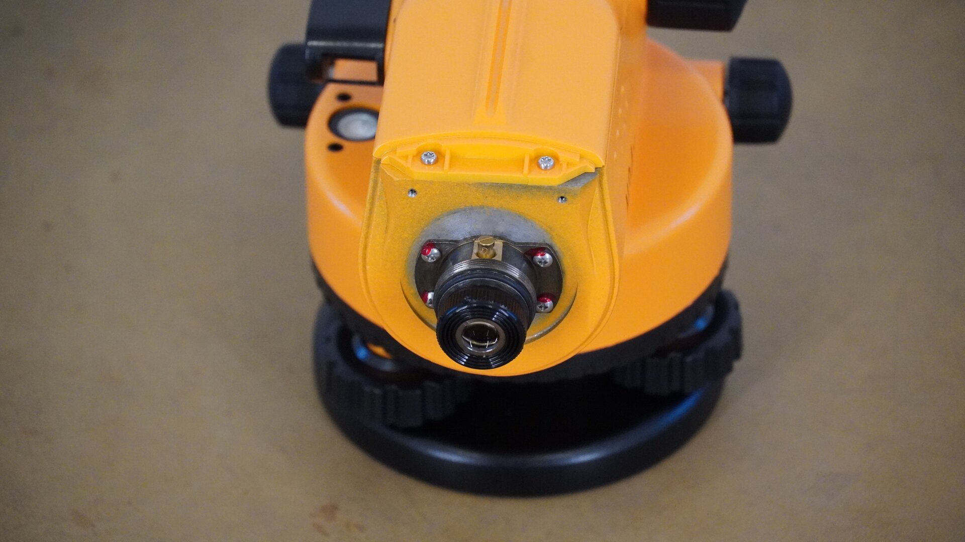 Zdjęcie ukazuje niwelator od strony okularu, z którego odkręcono obudowę. Widoczne są śruby mocujące okular do korpusu oraz czarne pokrętła po bokach i podziałka koła poziomego poniżej. Pod kołem poziomym znajdują się trzy śruby umieszczone na okrągłej podstawie, czyli spodarce.