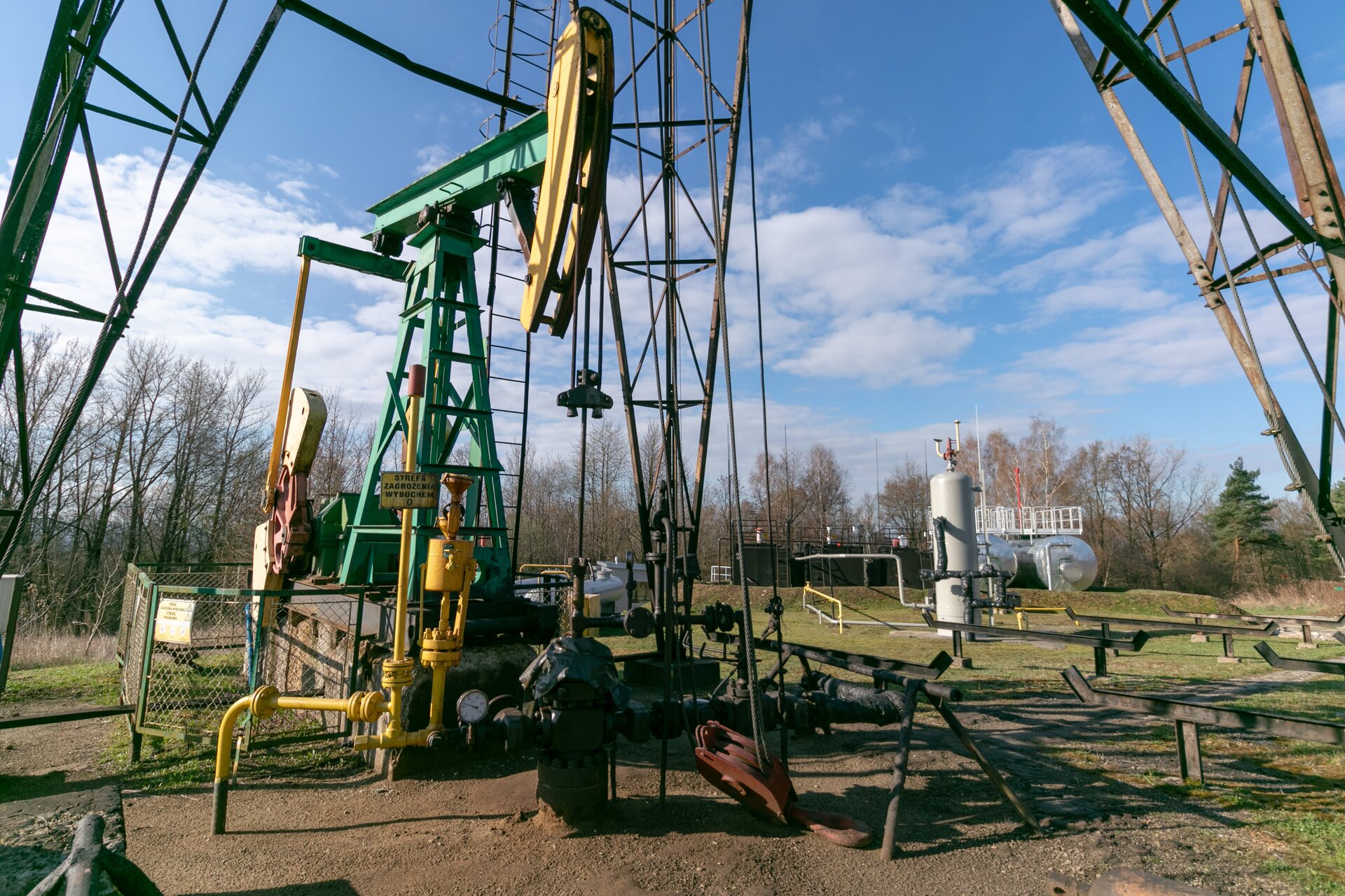 Na zdjęciu widać głowicę eksploatacyjną odwiertu pompowanego ropno‑gazowego wraz z przyległą armaturą do której zalicza się urządzenie pompowe typu IŻP-<math aria‑label=”dziewięć łamane na dwa tysiące”>10/1000 i rampą rurową. Głowica eksploatacyjna jest to urządzenie montowane na powierzchni terenu przy wylocie odwiertu, w celu jego zamknięcia. Na zdjęciu widać, że głowica to stalowe urządzenie które w całości przybrudzone jest na czarno. Podstawę urządzenia stanowi walcowaty kształt czyli kołnierz. Na nim znajduje się prwenter czyli urządzenie zabezpieczające. Prewenter jest w kształcie krzyża. Na prewentrze położona jest tkanina. Nad nim znajduje się trojak czyli rura w kształcie litery T umożliwiająca rozgałęzienie. Na górze urządzenia znajduje się laska pompowa w kształcie walcowatego pionowego pręta. Do końcówki trojaka, która jest skierowana w prawą stronę przyłączona jest armatura rurowa wraz z różnym typem zaworów. Przed głowicą znajduje się stalowy pomost. Do kołnierza głowicy przyłączone są po lewej stronie kolejne urządzenia rurowe wraz z zaworami. Nad jednym z zaworów znajduje się okrągłe urządzenie wskaźnikowe z białą tarczą i czarną wskazówką. Za nim znajduje się żółty element w kształcie cienkiego, długiego walca, który jest skierowany pionowo w górę. W punkcie stanowiącym około siedemdziesiąt procent wysokości znajduje się żółta tablica z czarnym tekstem brzmiącym: strefa zagrożenia wybuchem. Na końcu elementu znajduje się czerwony walec który jest nieznacznie szerszy od żółtego walcowatego elementu.Za głowicą znajduje się fragment indywidualnego żurawia pompowego, który wykorzystywany jest do wydobycia ropy naftowej z odwiertu. Żuraw jest stalową konstrukcją w kolorze zielono‑żółtym. U góry żurawia znajduje się stalowe zielone ramię zwane wahaczem. Na jego końcu, po prawej stronie znajduje się żółty łeb wahacza. Ramie z łbem jest w kształcie młotka. Środek wahacza osadzony jest na zielonej ramie nośne która jest w kształcie litery A. Lewy koniec wahacza przyłączony jest do tak zwanego podciągacza czyli dwóch stalowych pionowych belek przymocowanych do łuków osadzonych na przekładni. Za zieloną obudową przekładni widać fragment silnika. Żuraw pompowy jest wygrodzony niewielkim płotkiem. W oddali po prawej stronie ekranu widać separator odpowiedzialny za rozdział faz oraz znajdujące się za nim dwa srebrne zbiorniki cylindryczne i dwa zbiorniki skrzyniowe. Zbiorniki skrzyniowe są po lewej stronie zbiorników cylindrycznych. W tle widać drzewa.