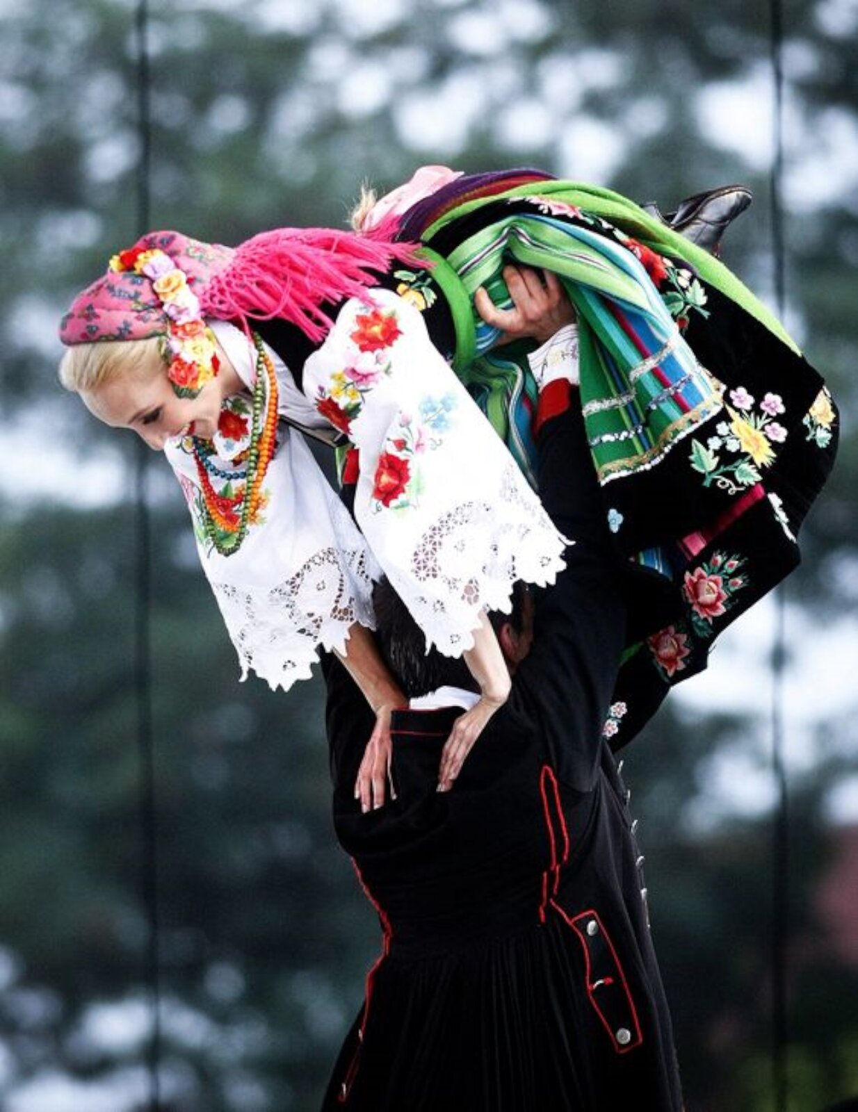 Zdjęcie przedstawia parę ubraną w strój ludowy - łowicki. Mężczyzna unosi kobietę nad głową, trzymając ją za biodra. Strój kobiety jest różnokolorowy, w kwiatowe wzory.