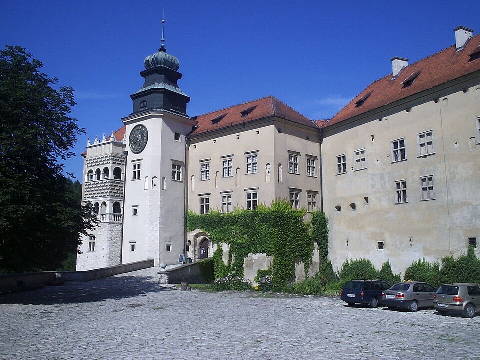Zdjęcie przedstawia murowany zamek z mostem wjazdowym, wieżę połączoną z zamkiem, czerwone dachy spadziste oraz brukowany podjazd.