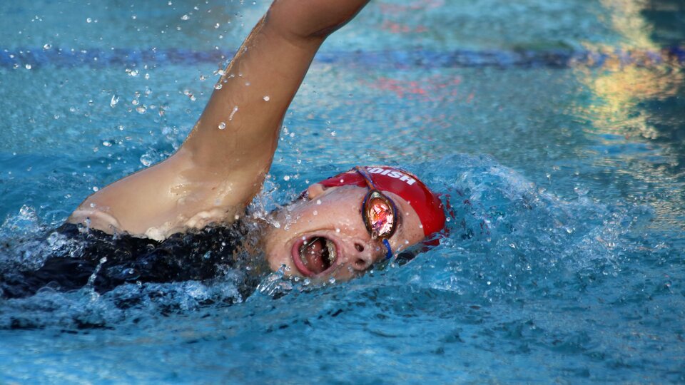 Zdjęcie przedstawia kobietę, który pływa w basenie. Ma uniesioną prawą rękę, nabiera powietrza, ma otwarte usta. Pływaczka ma na sobie strój kąpielowy, gogle oraz czepek.