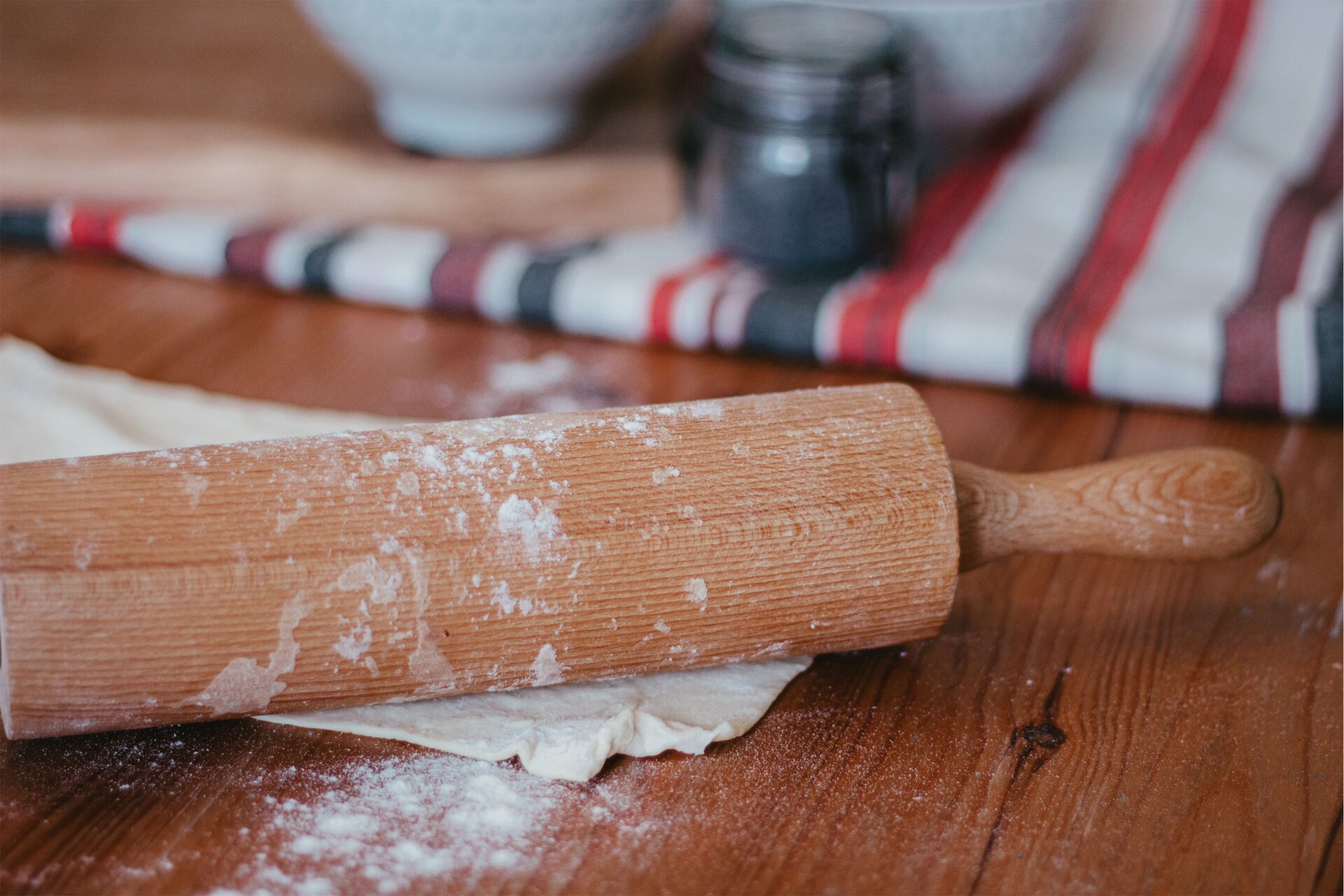 Zdjęcie przedstawia krok drugi formowania rogali świętomarcińskich. Ujęcie wykonane jest od góry pod skosem. Widoczny jest ciemnobrązowy drewniany blat na którym w lewej dolnej części grafiki znajduje się prostokątny płaski płat ciasta. Na nim ułożony jest drewniany jasnobrązowy wałek. W tle w górnej części zdjęcia dostrzec można niewyraźny zarys białej ścierki w czarne i czerwone pasy na której ustawiony jest przezroczysty słoik z niebieskim makiem. Obok dostrzec można jasnobrązową drewnianą deskę na której stoi mała biała miseczka.