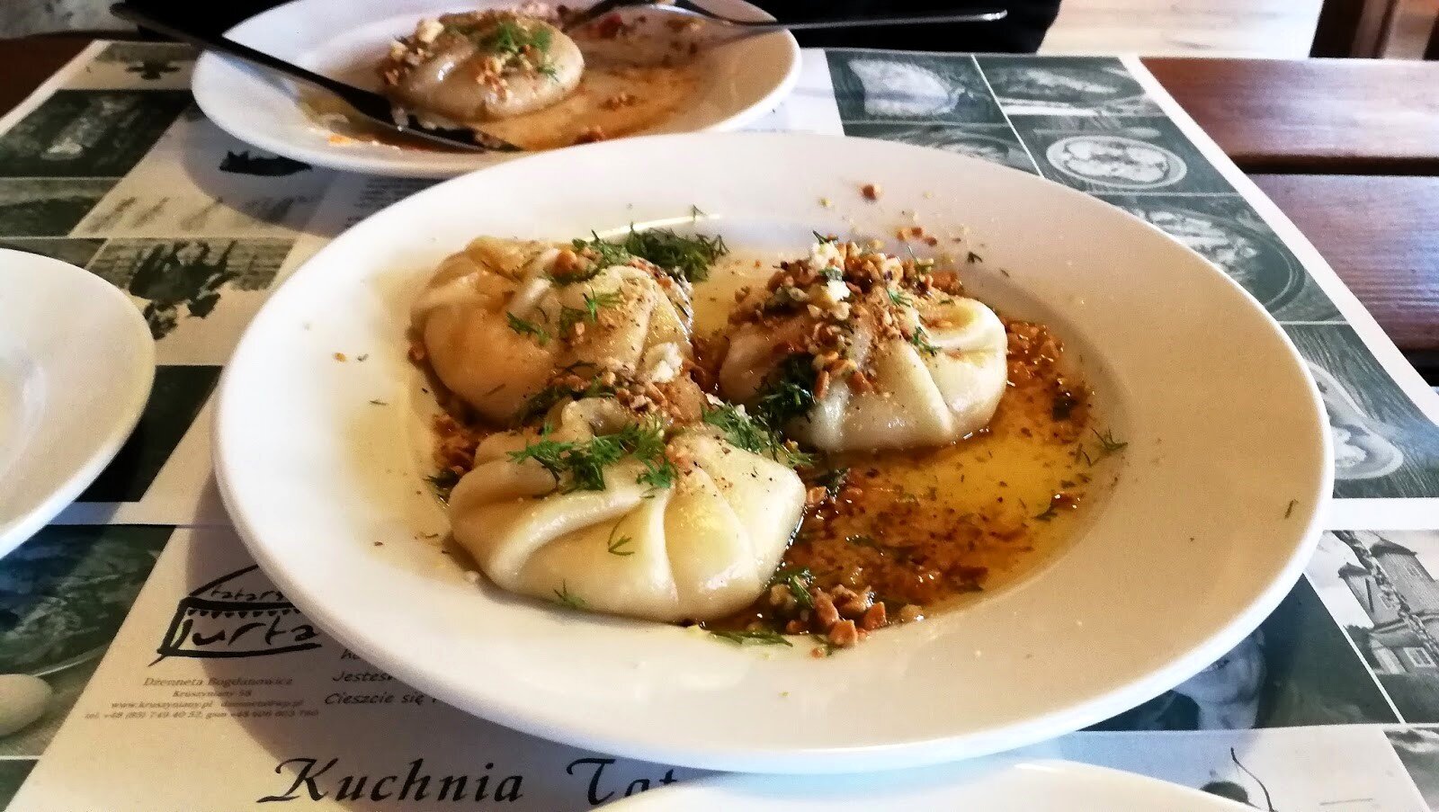 Zdjęcie przedstawia tatarskie danie podane w głębokim talerzu. Są to trzy pierożki w rosole, posypane koperkiem. Pierożki mają wygląd sakiewek. Na stole stoją jeszcze dwa inne talerze.