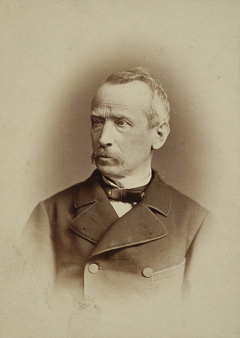 Zdjęcie przedstawia starszego mężczyznę. Ma krótkie, siwe włosy oraz gęste wąsy. Ma poziome zmarszczki na czole oraz zmarszczki wokół oczu. Ubrany jest w surdut.