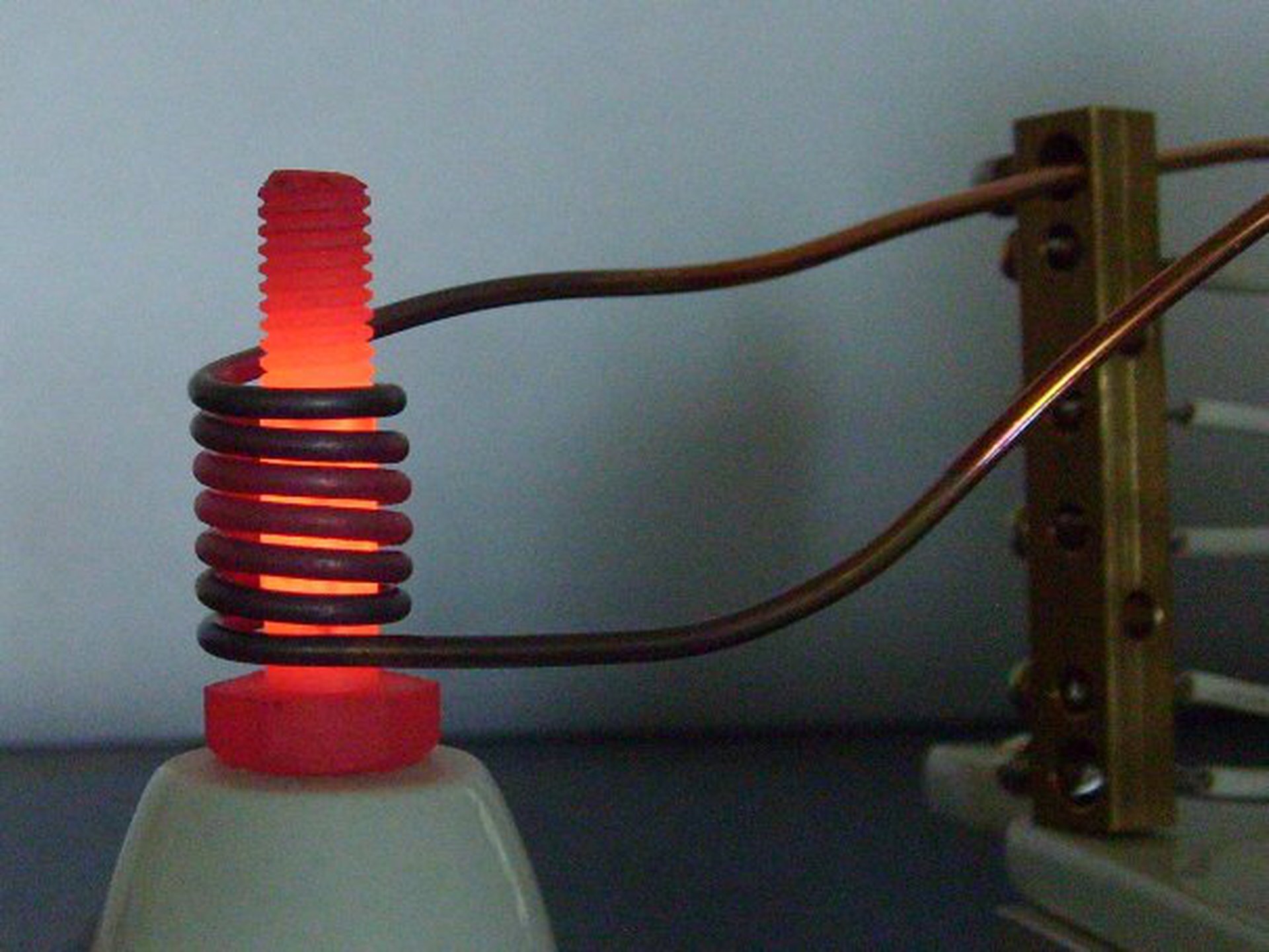 Rys. 2. Zdjęcie przedstawia metalowy wkręt ustawiony pionowo na łebku. Wkręt znajduje się wewnątrz przewodu elektrycznego, zwiniętego w linię śrubową. Dolny i górny koniec przewodu poprowadzony jest w prawo do źródła prądu zmiennego, znajdującego się domyślnie poza fotografią. Wkręt rozgrzany jest do czerwoności.