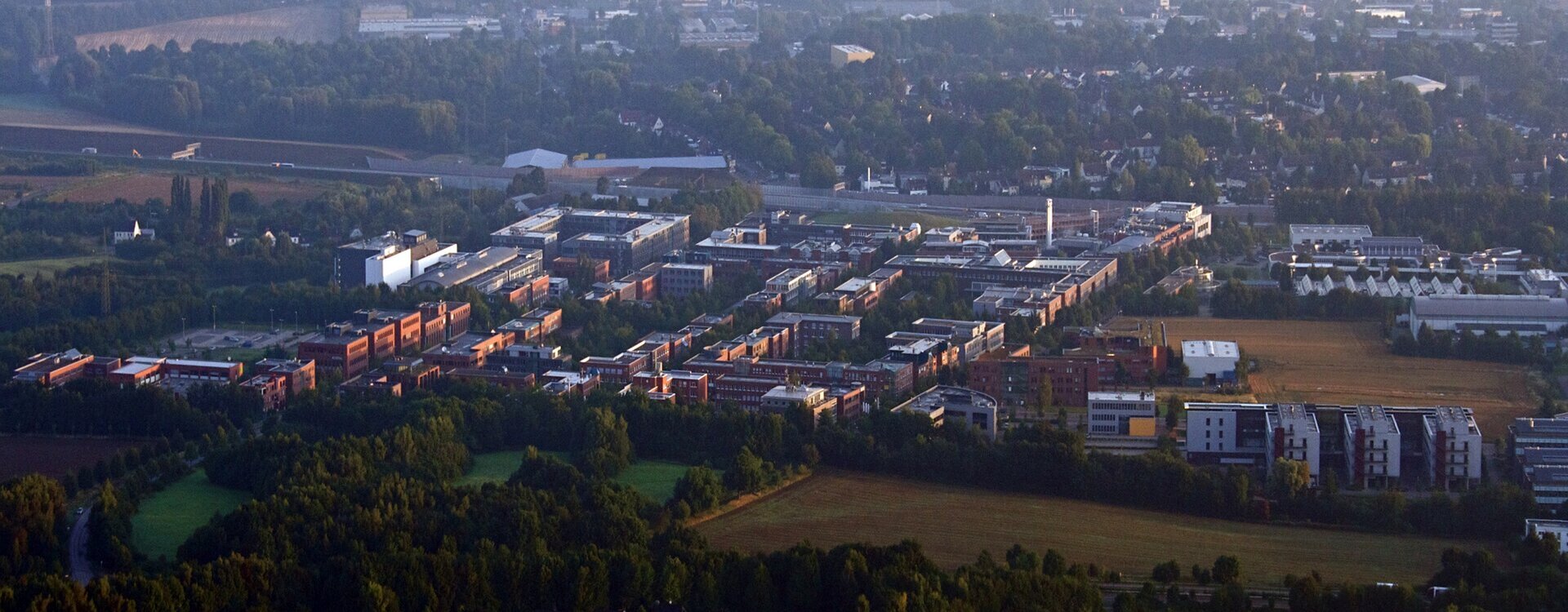 Zdjęcie lotnicze kompleksu kilkupiętrowych budynków biurowych pod miastem. Za nimi osiedle domów pośród drzew. Przed nimi pola i lasy.