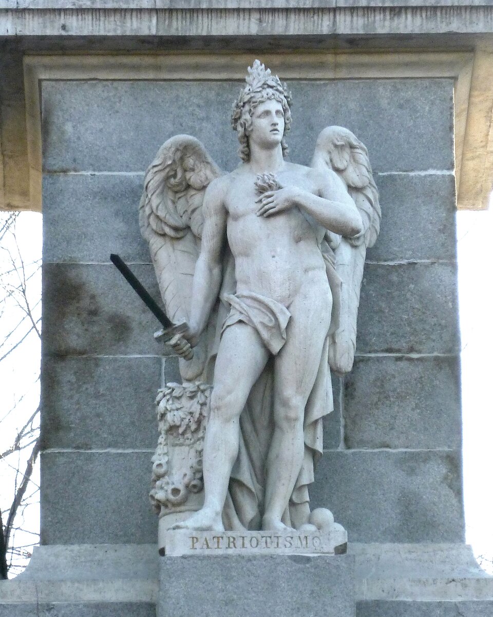 Zdjęcie przedstawia rzeźbę mężczyzny stojącą na tle muru budynku. Rzeźba przedstawia młodego, nagiego mężczyznę ze skrzydłami, który trzyma w prawej ręce uniesiony miecz. Mężczyzna jest dobrze zbudowany, ma muskuły i umięśnione nogi. Przyrodzenie zakrywa fragment szaty, która ułada się miękko za plecami. Głowę okala wieniec laurowy. Lewa ręka mężczyzny spoczywa na piersiach, w okolicy serca. Pod rzeźbą widnieje łaciński napis, który w tłumaczeniu na język polski brzmi: patriotyzm.