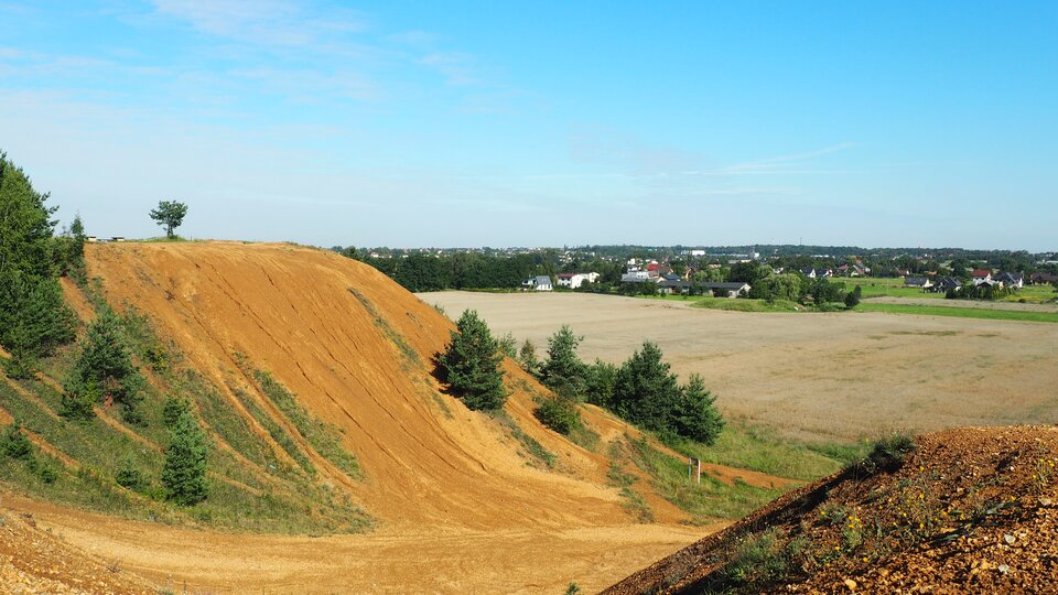 Zdjęcie przedstawia wzniesienie. Gleba ma na nim kolor pomarańczowy. Gdzieniegdzie na wzniesieniu rosną drzewa. W tle jest pole, a daje zabudowania. 