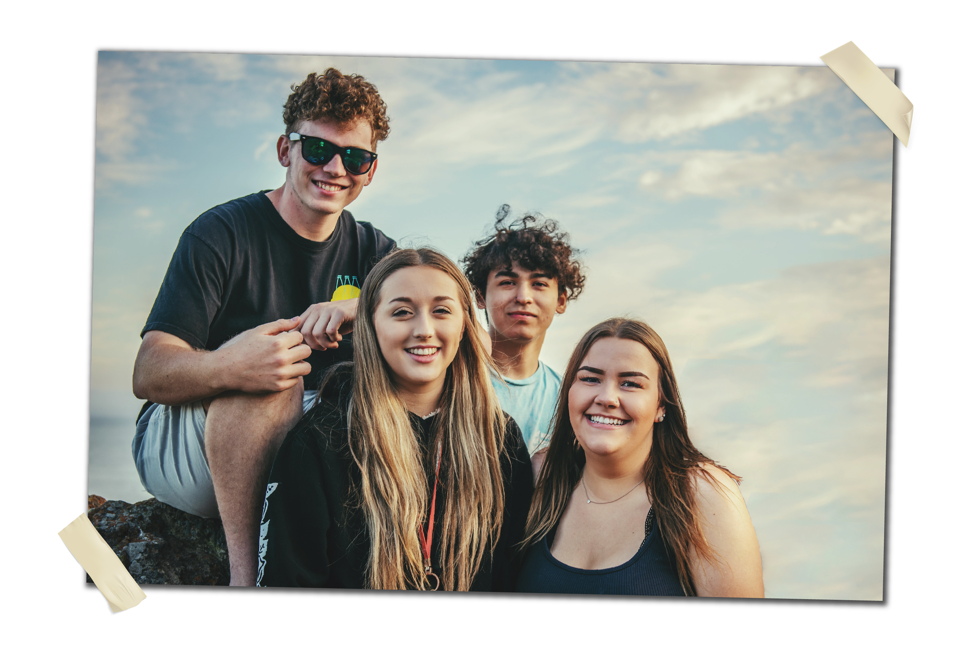 Zdjęcie przedstawia grupę młodych ludzi, dwóch chłopców i dwie dziewczyny. Są uśmiechnięci, ubrani w sportowe stroje. Siedzą na kamiennym podłożu, w tle widoczne niebo z chmurami.