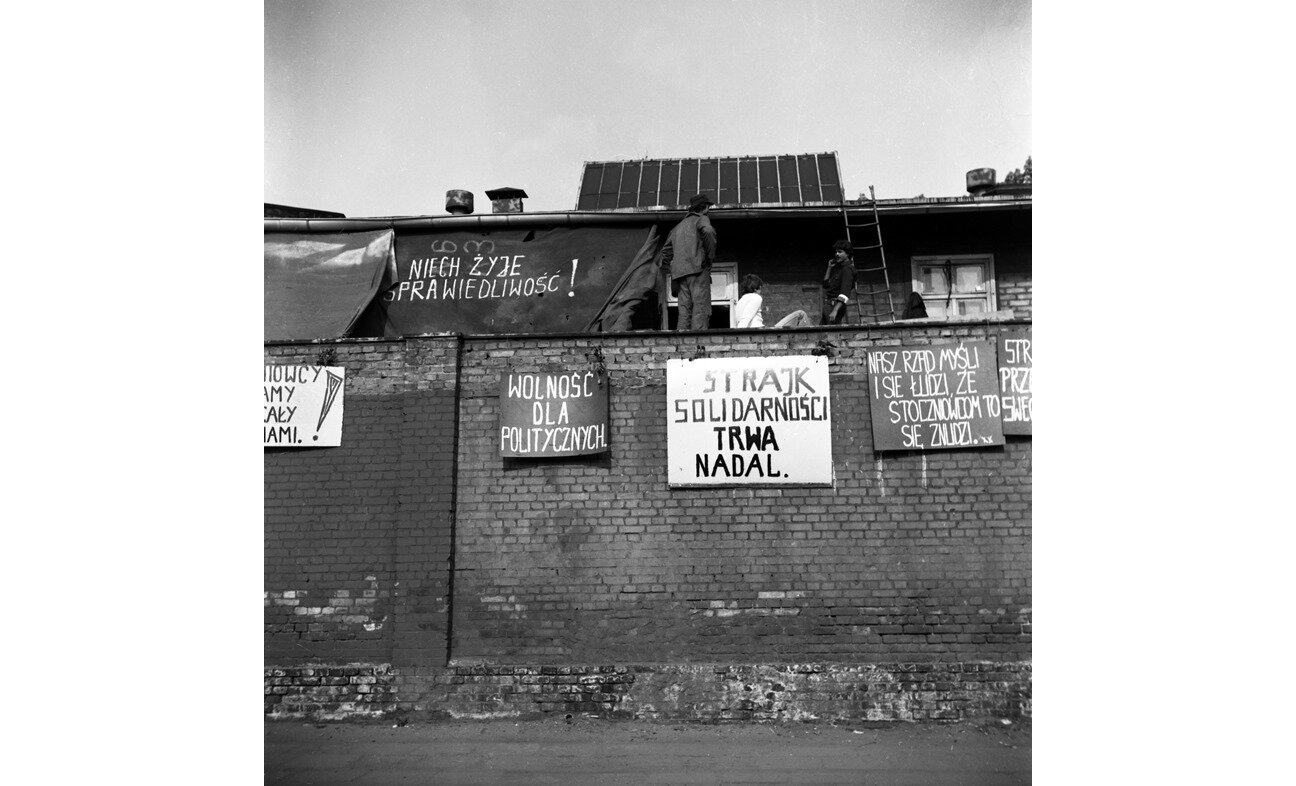 Czarno-biała fotografia przedstawiająca transparenty wywieszone na murze stoczni w Gdańsku. Znajdują się na nich hasła "niech żyje sprawiedliwość", "wolność dla politycznych", "strajk solidarności nadal trwa", "nasz rząd myśli i się łudzi, że stoczniowcom to się znudzi".