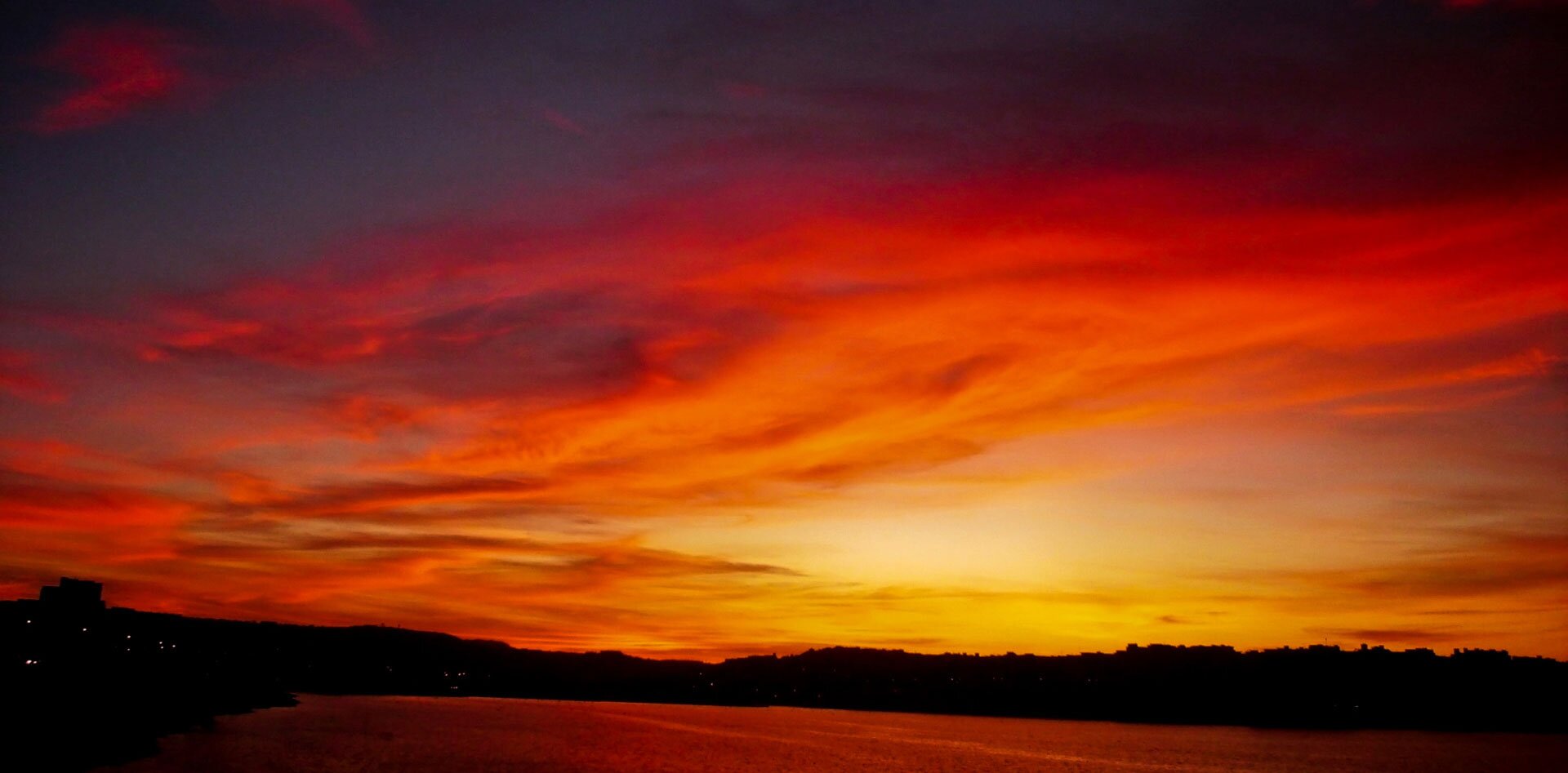 Fotografia przedstawia zachód słońca nad zbiornikiem wodnym. Niebo jest czerwone, miejscami pomarańczowe, z zaznaczonymi wyraźnie chmurami