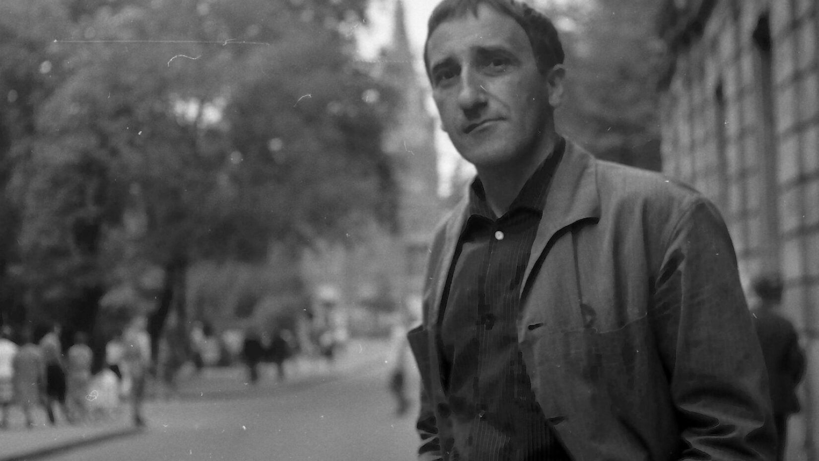 Fotografia przedstawia Tadeusza Kantora, który został sportretowany na tle tętniącego życiem miasta. Kantor to mężczyzna w średnim wieku, ma on krótkie, proste włosy opadające na czoło, zarysowane cienie pod oczami i zmarszczki mimiczne przy ustach. Ubrany jest w szarą marynarkę i ciemną koszulę w paski. Kantor pozuje z półprofilu i z lekkim uśmiechem spogląda w obiektyw.