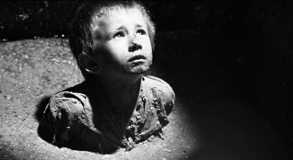 Zdjęcie przedstawia małego chłopca zanurzonego po ramiona w odpadkach. Dziecko patrzy się z rozpaczą do góry.