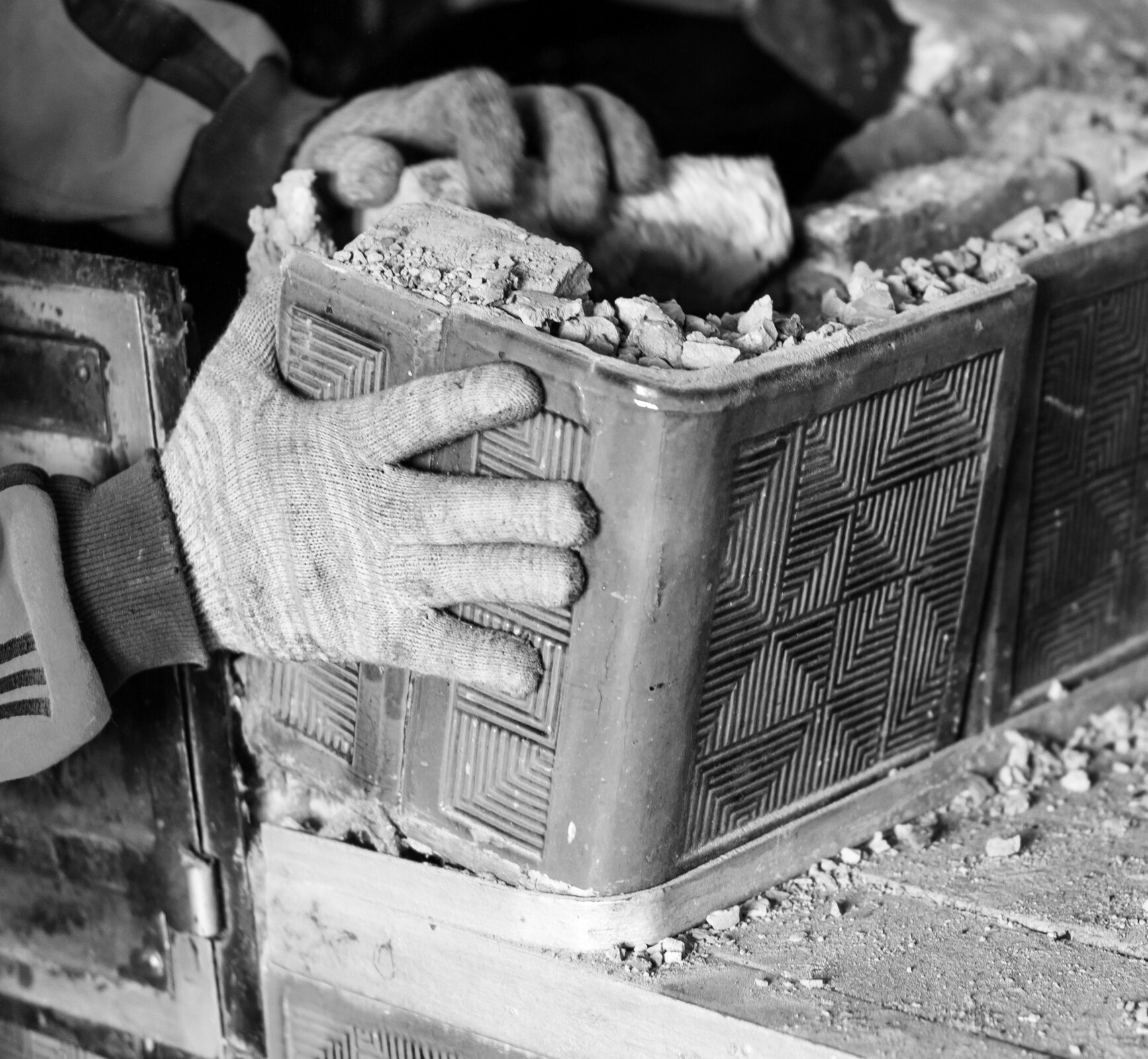 Fotografia przedstawia dwie dłonie w rękawiczkach demontujące narożny kafel w piecu kaflowym. Kafel zdobiony jest ornamentami w kształcie rombów.