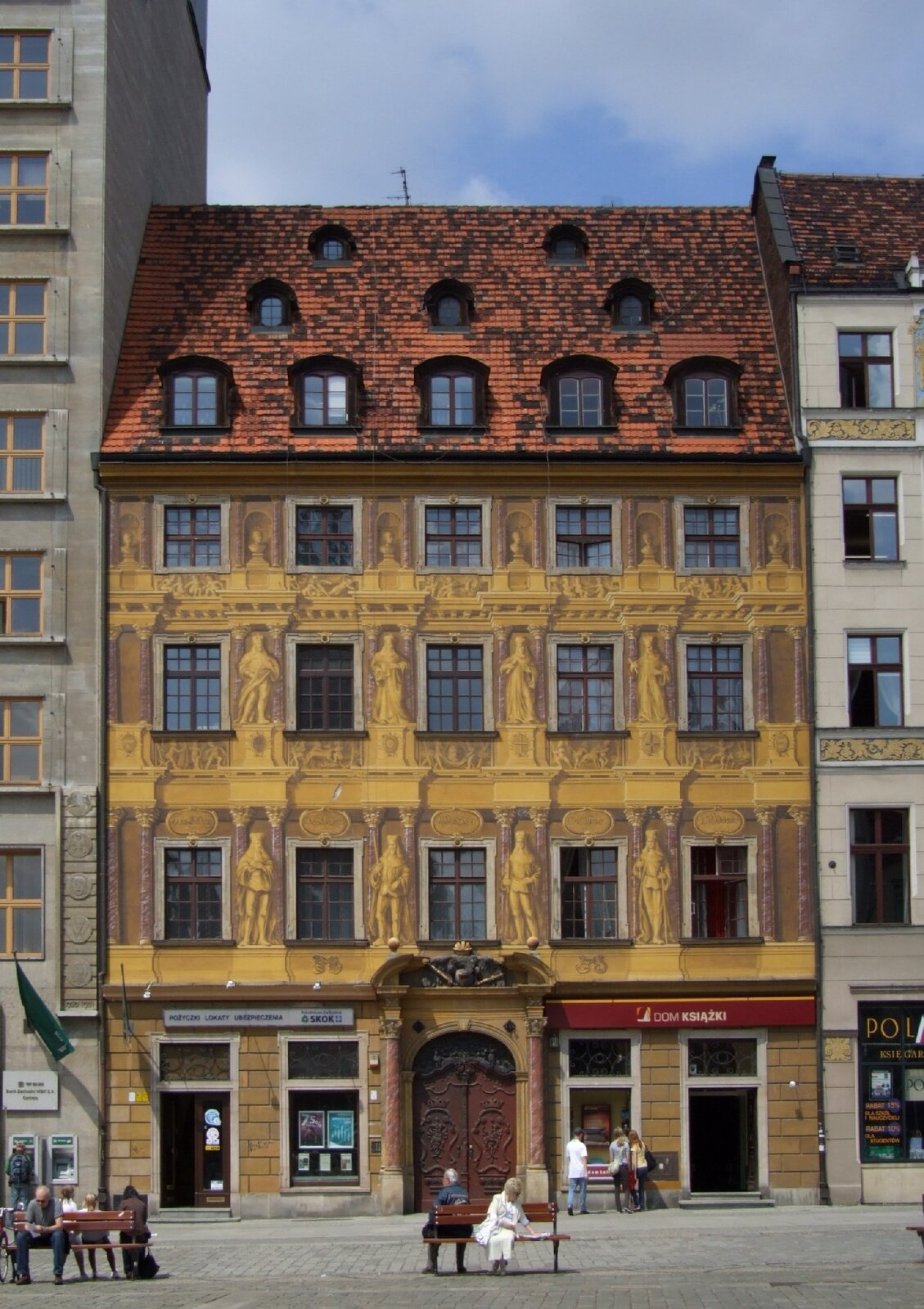 Zdjęcie przedstawia kamienicę. Elewacja budynku jest żółta, a na niej znajdują się liczne, brązowe zdobienia. Kamienica składa się z czterech pięter, a także skośnego, czerwonego dachu, w którym umieszczone zostały okna sugerujące trzy dodatkowe piętra. Kamienica znajduje się pomiędzy dwoma jasnymi budynkami. Przed kamienicą wybetonowana droga, po której chodzą przechodnie.