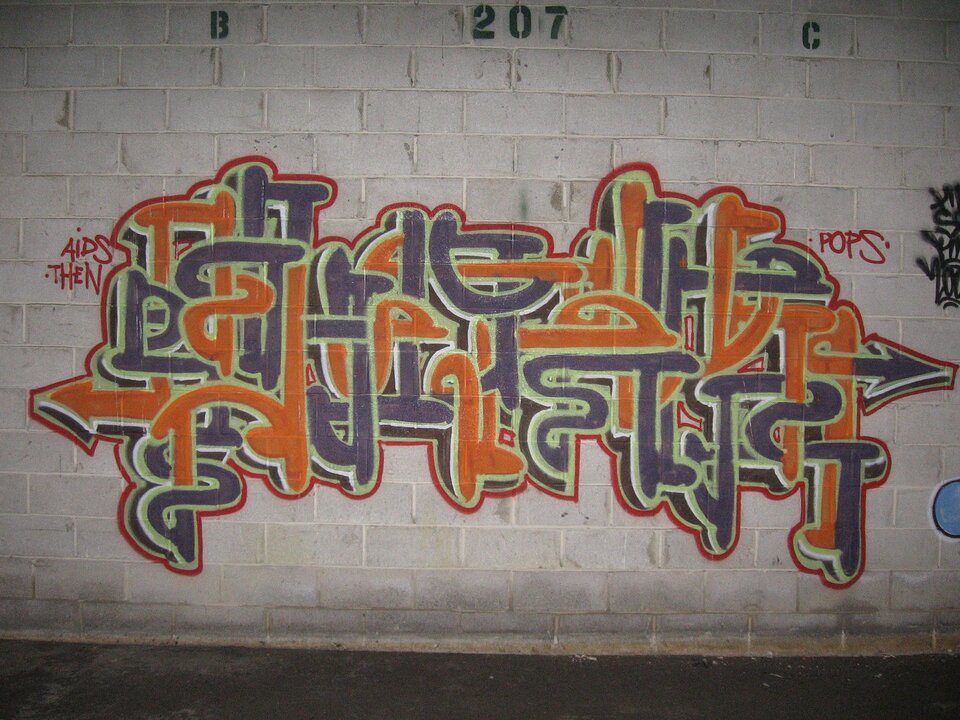 Zdjęcie przedstawia graffiti namalowane na jasnej ścianie. Ściana jest koloru białego. Graffiti namalowane jest w kolorze szarym oraz pomarańczowym.