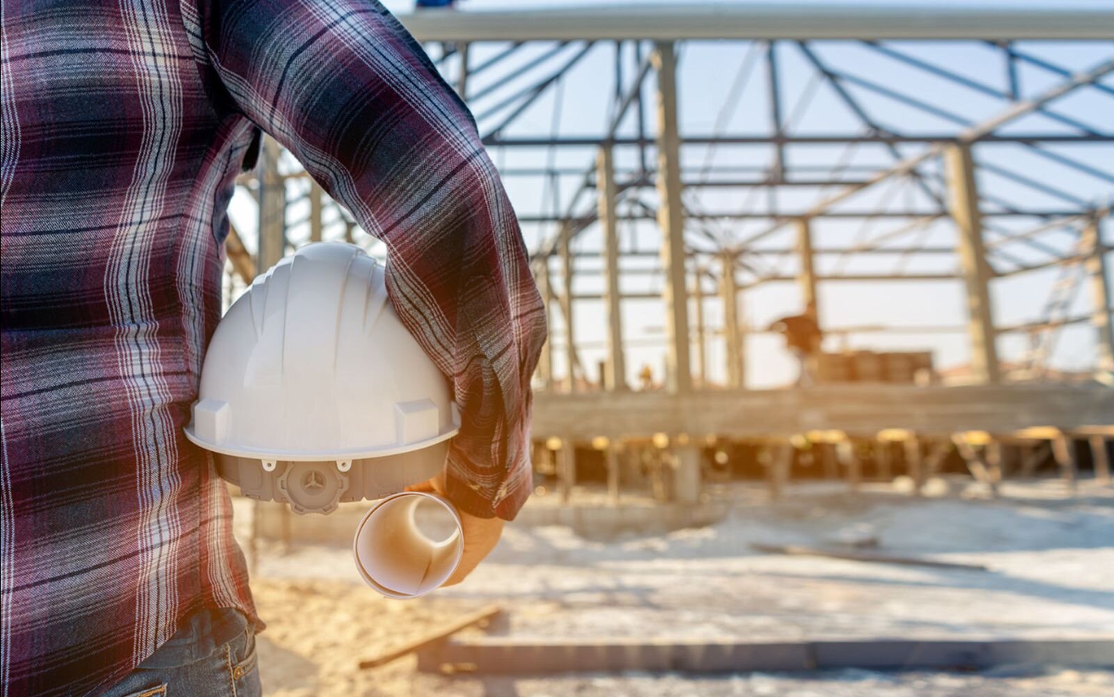 Na ilustracji z lewej strony znajduje się stojący tyłem człowiek. Trzyma w ręku kask oraz papierowy rulon. Tło stanowi wielkich rozmiarów konstrukcja budowlana. Fotografia przedstawia fragment terenu budowy.