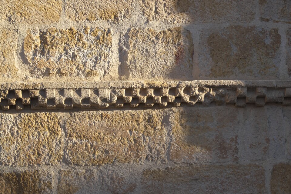 Detal romańskiej ściany z kamienia: poziomy fryz z drobnych, regularnych kostek tworzących ząbkowany pas dekoracyjny, osadzony między surowymi blokami muru.