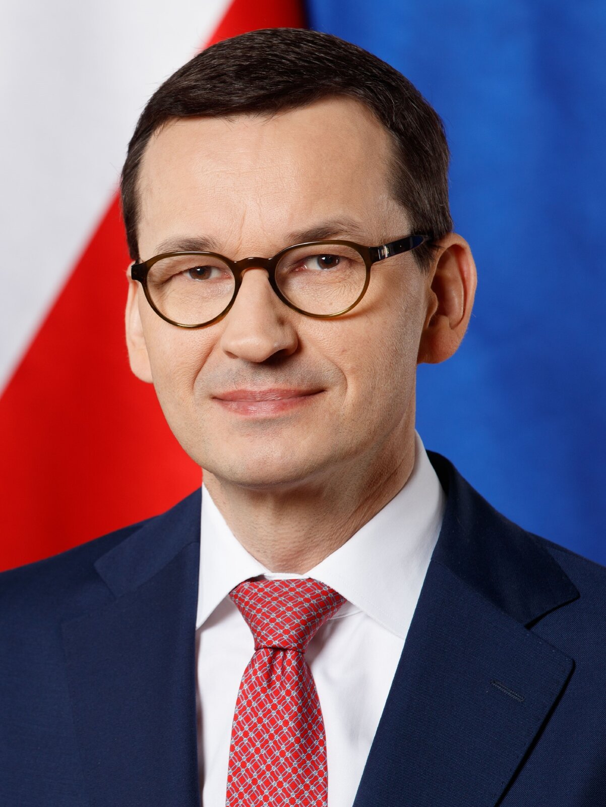 Na zdjęciu premier Mateusz Morawiecki, ubrany w garnitur, koszulę i krawat. Ma na sobie okulary. W tle z lewej strony znajduje się fragment flagi Polski, z prawej strony fragment flagi Unii Europejskiej.
