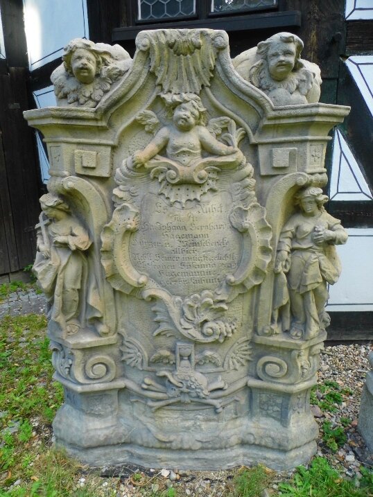 Zdjęcie przedstawia zabytkowe epitafium z XVIII wieku. Jest wykonane z kamienia, bogato dekorowane rzeźbami postaci, głowami aniołków, wolutami, gzymsami i płaskorzeźbami ustawionymi symetrycznie po obu stronach nieregularnej tablicy z inskrypcją, trzymanej przez aniołka. Całość wygląda jak pomniejszony fragment ściany barokowego budynku. 