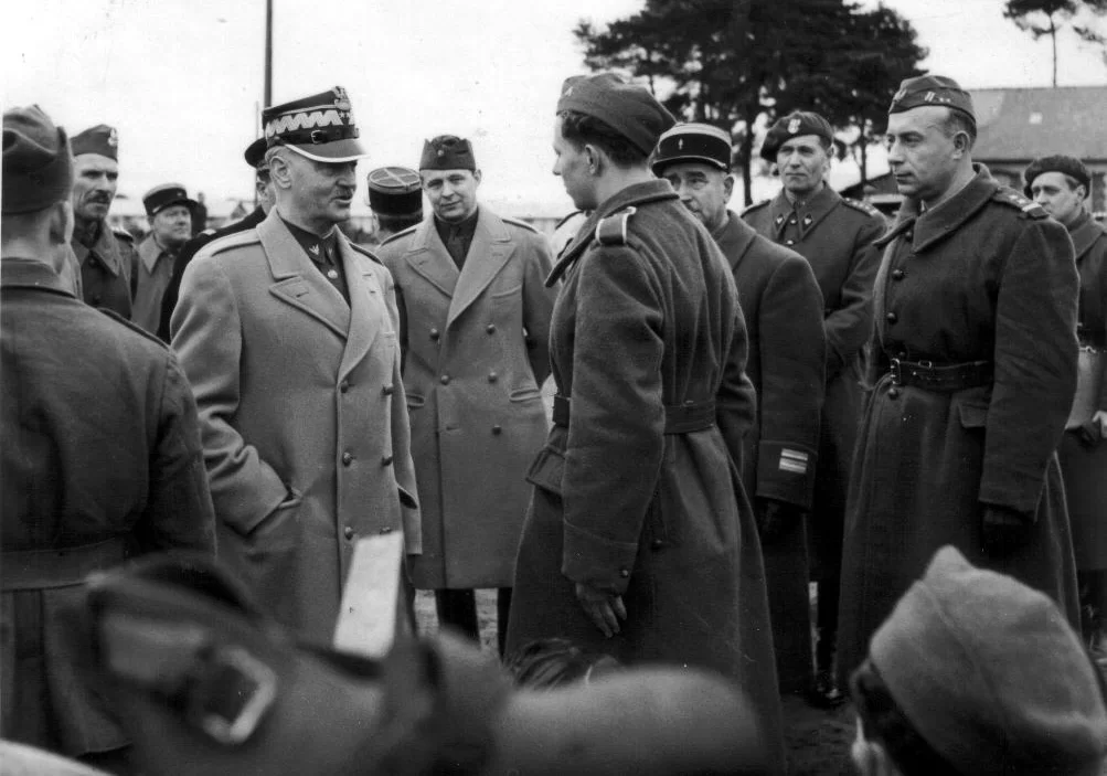 Kliknij, aby powiększyć Zdjęcie. Mężczyzna w wojskowym płaszczu i czapce stoi na przeciwko żołnierza. Patrzą na siebie i rozmawiają. Wokół nich kilku żołnierzy, wszyscy na nich patrzą. W tle dach zabudowania, kilka koron drzew.