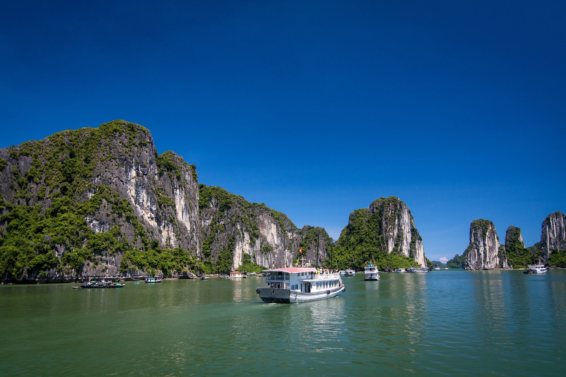  Na pierwszym planie znajduje się nieduży statek turystyczny płynący po wodzie. Obok niego są  mniejsze statki i łódki. Po lewej stronie widać wysokie, pionowe skały porośnięte zielenią, które znajdują się wzdłuż brzegu, a w oddali tworzą wraz ze skałami formację na kształt bramy. W tle czyste, błękitne niebo.