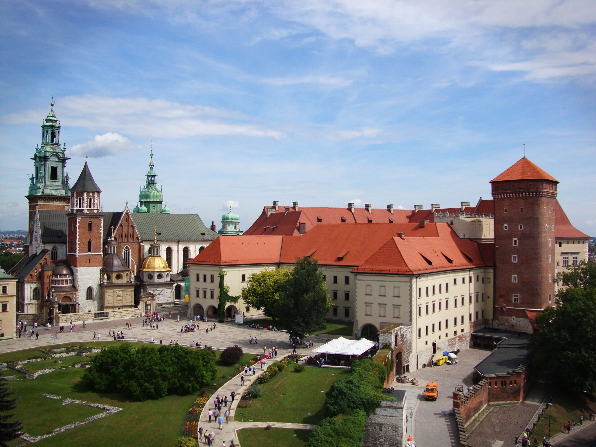 Fotografia przedstawiająca zamek i katedrę na Wawelu. Przed nimi plac, na którym zgormadzeni są mieszkańcy i turyści. Z przodu trawa i krzewy. W tle miasto.
