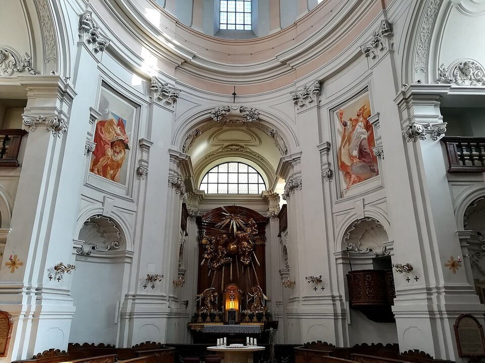 Ilustracja przedstawia wnętrze kościoła Świętej Trójcy w Salzburgu. Wnętrze jest w całości tynkowane na biało. Jego dekoracja ograniczona jest przede wszystkim do sztukaterii podkreślającej detal architektoniczny. Jedynie, poniżej kopuły, na przeciwległych ścianach, znajdują się dwa freski w barwach czerwonych, białych i żółtych. W oddali widać ołtarz, ściana za ołtarzem wykonana jest z ciemnego drewna, umieszczone są na niej wyrzeźbione postacie.