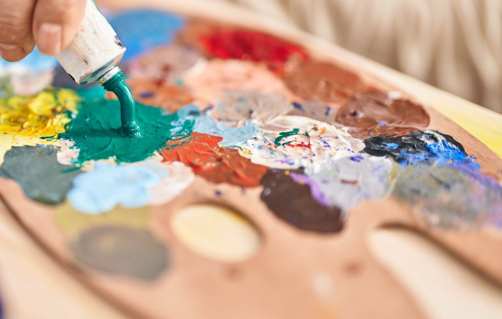 Zdjęcie przedstawia paletę malarską z wyciśniętymi farbami. Wyraźna jest tylko środkową część palety, gdzie ręka wyciska z tubki farbę. Pozostałe części są niewyraźne.