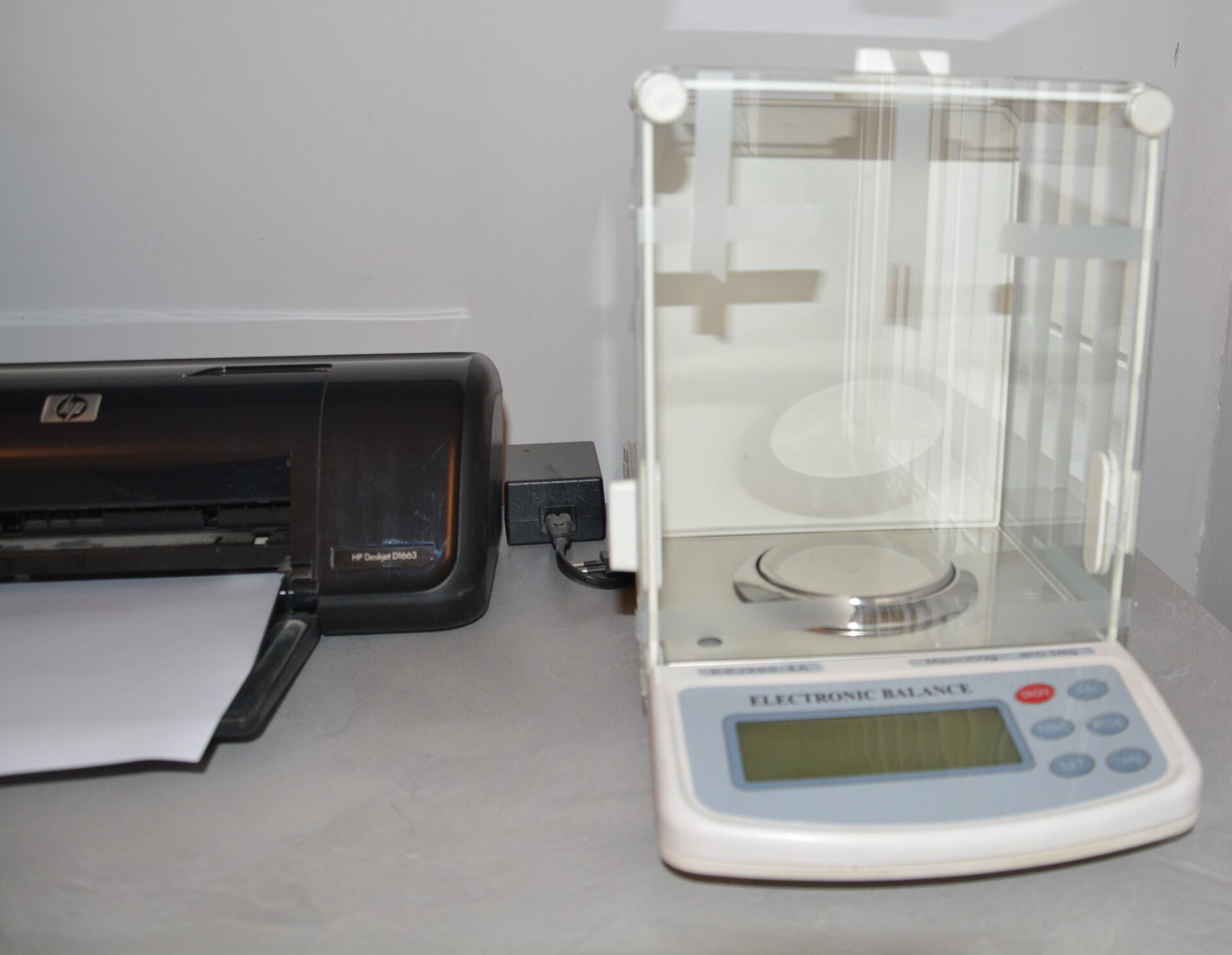 Zdjęcie przedstawia wagosuszarkę. Po prawej stronie widoczne jest urządzenie złożone z podstawy, na której widoczny jest prostokątny wyświetlacz oraz sześć przycisków, w tym jeden czerwony. Na podstawie znajduje się przeszklony prostopadłościan, w którego dolnej części znajduje się niski metalowy walec. Po lewej stronie widoczna jest drukarka z kartką włożoną do podajnika.