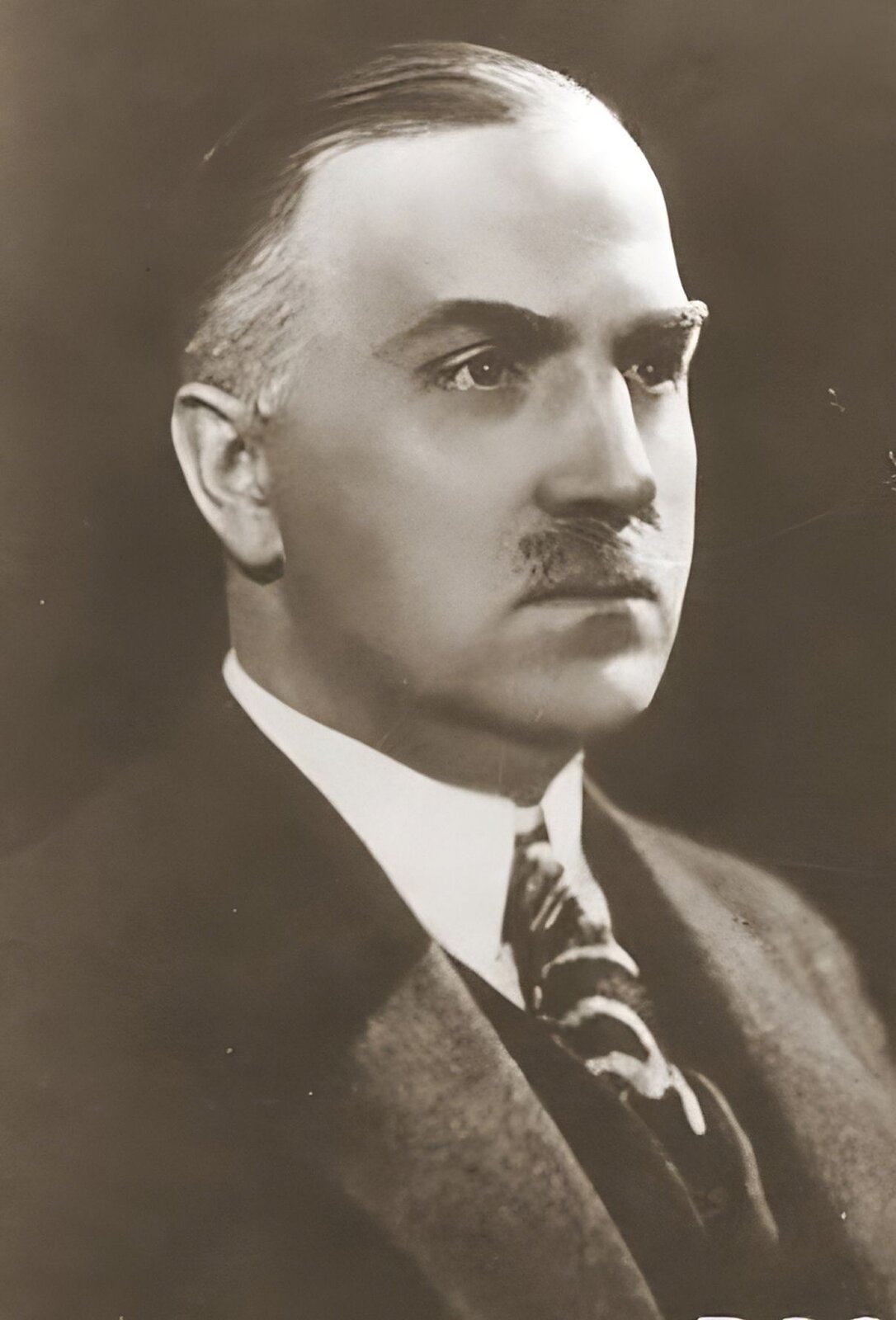 Kliknij, aby powiększyć. Fotografia przedstawia portret mężczyzny w średnim wieku. Ma ciemne włosy, po bo bokach siwiejące, zaczesane do góry. Gęste, brwi, prosty nos i małe wąsy. Ubrany jest w białą koszulę, ciemną marynarkę i krawat we wzory. Tło jest ciemne jednolite.