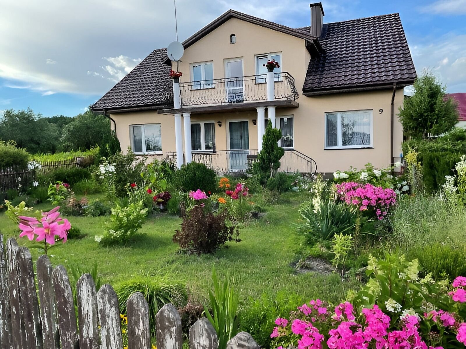 Fotografia przedstawia dom jednorodzinny z brązowym dachem i beżową elewacją. Wokół trawa i kępy różowych kwiatów, Dom ogrodzony jest drewnianym płotem. 