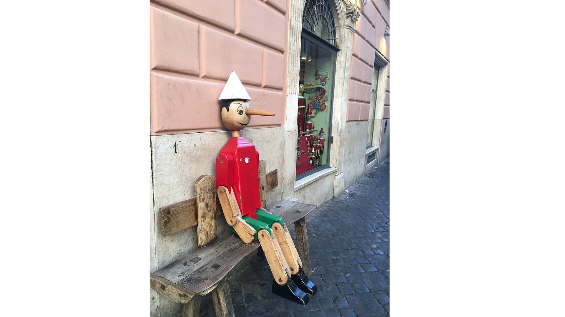 Zdjęcie przedstawia znaną z książki, bajek i filmów drewnianą kukiełkę o imieniu Pinokio. Pinokio to postać chłopca, cała wykonana z drewna, ma charakterystyczny długi nos. Na zdjęciu Pinokio siedzi na ławce stojącej na ulicy, przy której stoją stare kamienice. 