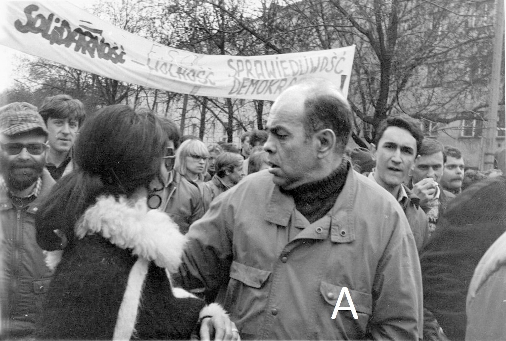 Zdjęcie przedstawia postawnego, dojrzałego mężczyznę. Ukazany jest jego lewy profil. Mężczyzna jest łysiejący, ma niewielki, zadarty nos. Ubrany jest w golf i kurtkę. Mężczyzna idzie pod rękę z kobietą w kożuchu. Za ich plecami idzie tłum ludzi. Widać transparent z napisem Solidarność Wolność Sprawiedliwość Demokracja.