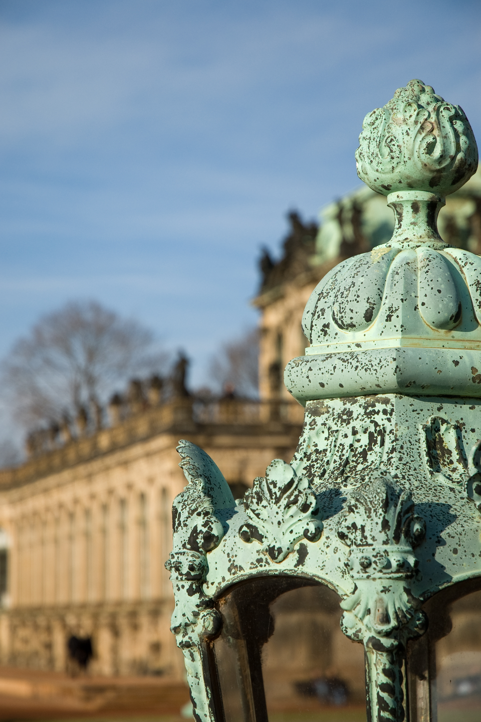 Na zdjęciu widoczny jest fragment zdobionej mosiężnej lampy pokrytej zielonkawoniebieskim nalotem patyny. W tle za lampą widoczne są zabytkowe zabudowania pałacowe.