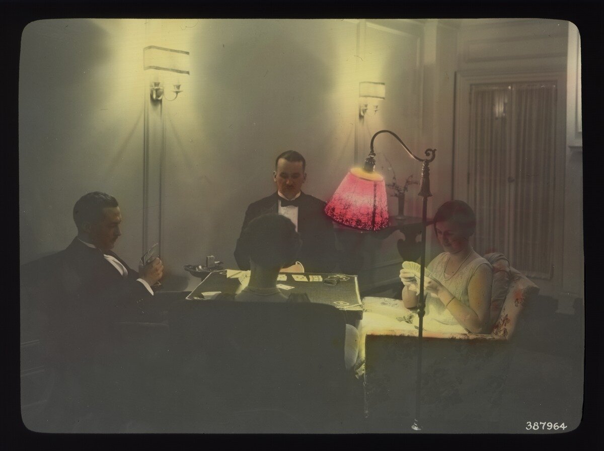 Stare zdjęcie przedstawiające czwórkę osób grających w pokera, które siedzą przy stole w jasnym pomieszczeniu. Przy czarnym stole siedzą dwie kobiety ubrane w sukienki oraz dwóch mężczyzn ubranych w smokingi. Stół oświetla lampa z różowym kloszem, która stoi pomiędzy dwoma siedzeniami.