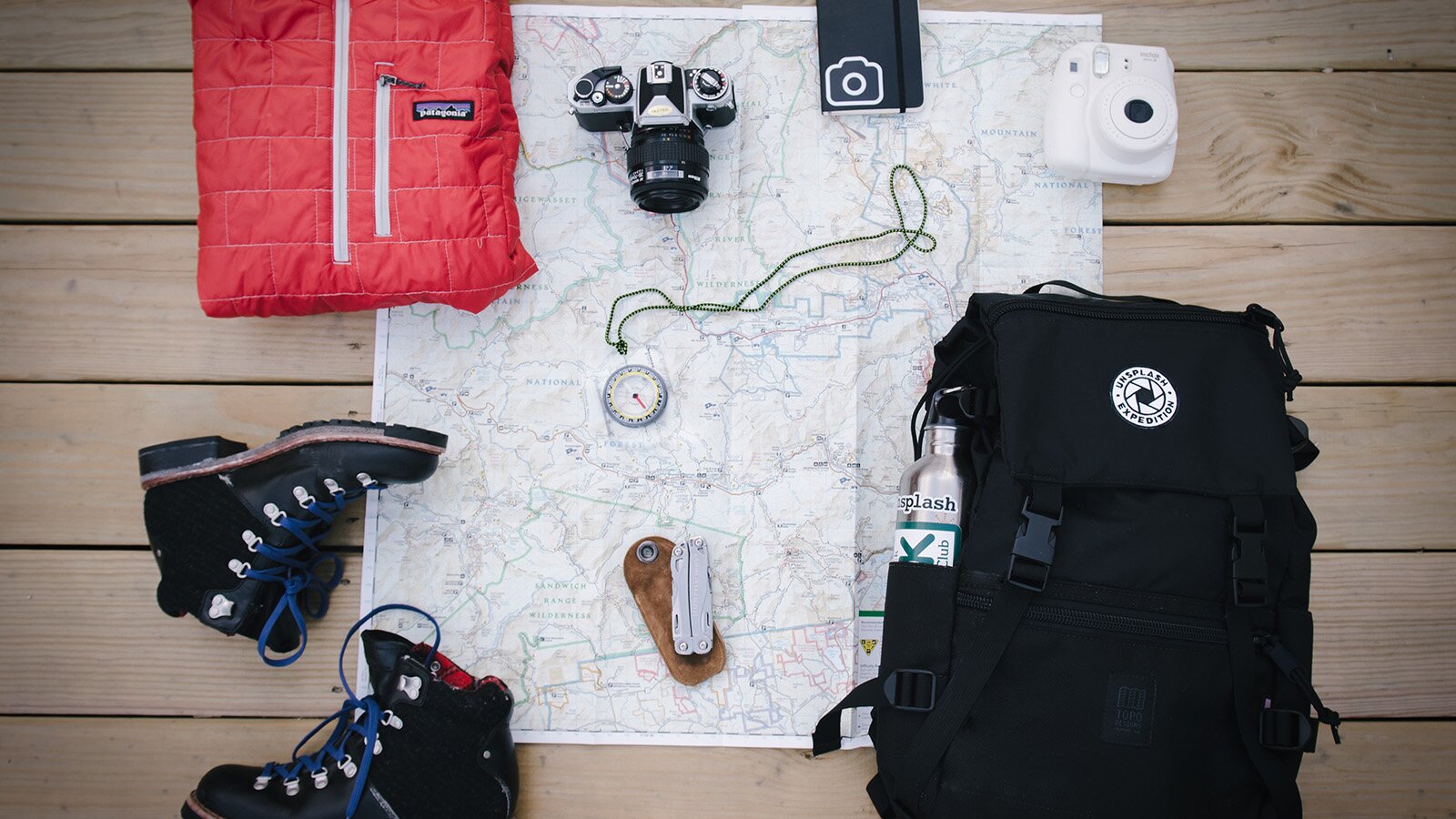 Zdjęcie przedstawia rozłożoną mapę, a na niej ułożone przedmioty przydatne w wędrówkach górskich. Wokoło mapy oraz na niej ułożono: złożoną kurtkę, obok której leży aparat, kompas, notes, pod nimi są buty trekkingowe oraz plecak. 