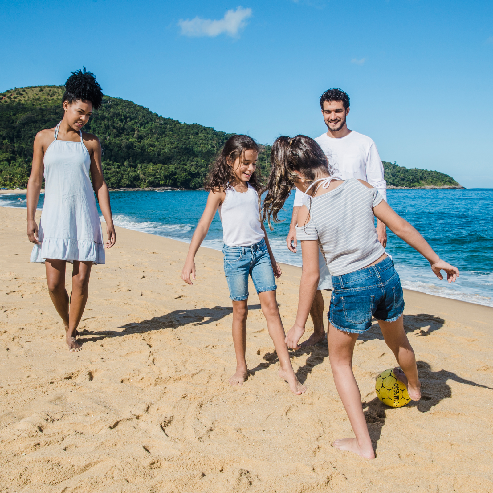 Zdjęcie przedstawia kobietę, mężczyznę i dwie dziewczynki w wieku szkolnym, grających w piłkę na plaży.